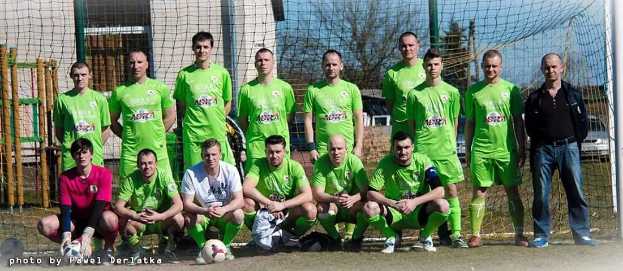 Gruppe von Männern in grünen T-Shirts auf einem Fußballfeld mit einem Ball davor, neben einem Torpfosten, mit Bäumen, Gebäuden und Fahrzeugen im Hintergrund.