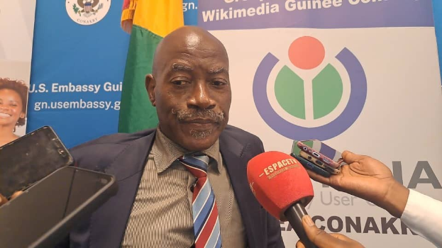 Ein Mann im Anzug spricht mit Reportern, während ein Mikrofon und ein Aufzeichnungsgerät im Vordergrund zu sehen sind, und Banner, Flaggen und eine Frau im Hintergrund.