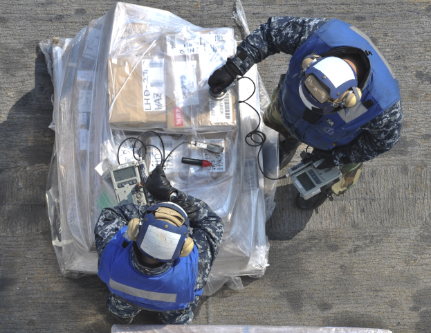 Zwei Männer in blauen Uniformen und Helmen arbeiten an einem großen Drohnenmodell und halten verschiedene Gegenstände in den Händen, während eine Plastikabdeckung vor ihnen auf dem Boden liegt.