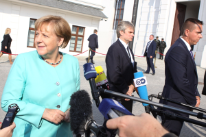 Bundeskanzlerin Angela Merkel spricht vor dem Kanzleramt in Berlin mit Reportern, umgeben von einer Gruppe von Menschen, einige halten Mikrofone, mit Gebäuden im Hintergrund.