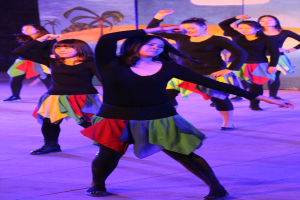 Gruppe junger Frauen in bunten Röcken und Schuhen tanzt energisch auf einer Bühne mit einem Gemälde im Hintergrund.