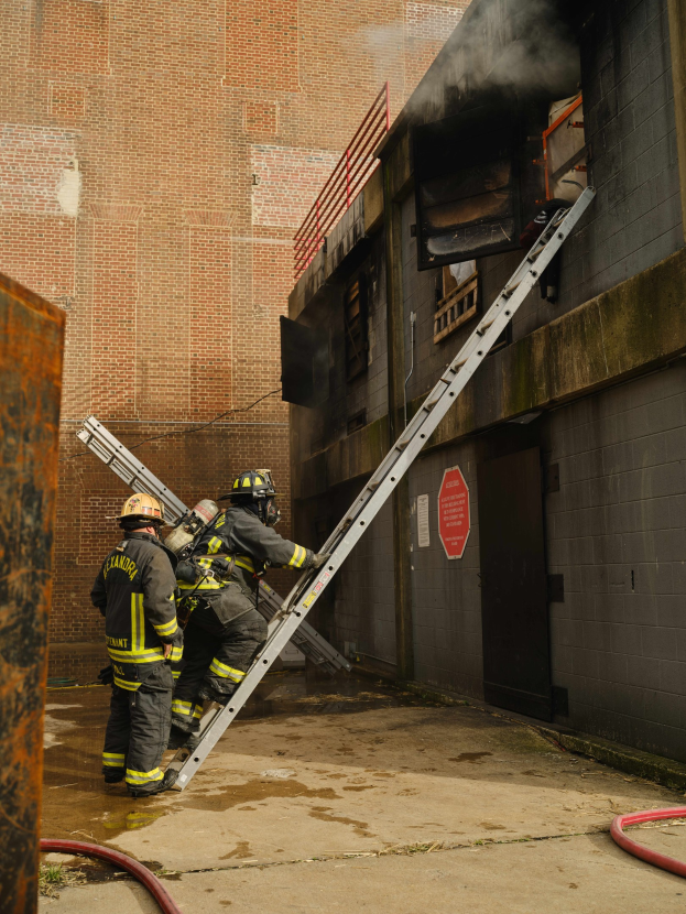 Feuerwehrleute mit Helmen klettern an einer Leiter zu einem raucherfüllten Gebäude hoch, während ein metallisches Objekt und Bodenschläuche zu sehen sind.