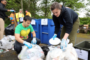 Menschen im Park Müll auf Tellern sammeln, mit Abfall wie Plastikdeckel und Flaschen auf dem Boden, ein Mülleimer und eine Bank rechts daneben und Trees und einen klaren Himmel im Hintergrund.