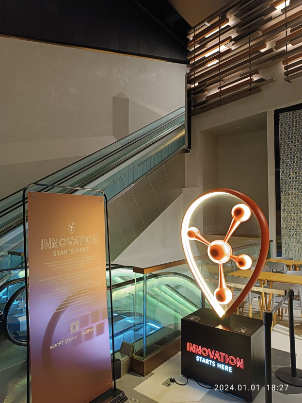 Großes Schild in einem Gebäude mit einer Rolltreppe im Hintergrund, mit Tischen und Stühlen auf der rechten Seite und einer Tafel mit "Innovation Starts Here" auf der linken Seite, was darauf hinweist, dass es Teil der HSTP20 Expo ist.