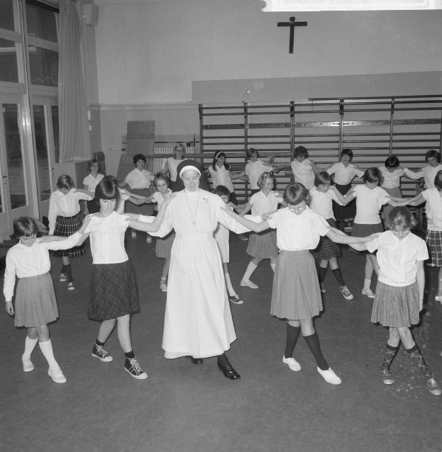 Schwarzes und weißes Bild von Kindern in Schuluniformen, die in einem Raum mit einer Wand mit einem Wandkreuz, Glastüren mit Vorhängen und Deckenlampen tanzen.