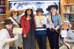 Eine Gruppe von Frauen steht nebeneinander vor Bücherregalen, jede hält ein "Women's Vote"-Plakat, einige tragen Hüte und ein Bildschirm ist im Hintergrund zu sehen.