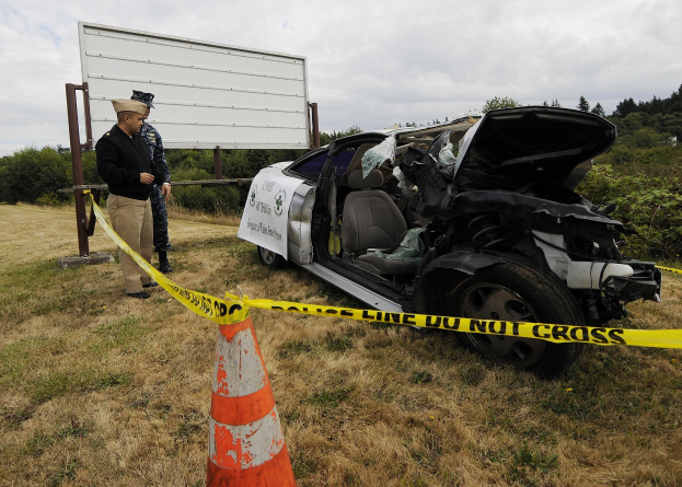 Polizist in Uniform inspiziert ein schwer beschädigt Auto mit eingedrückter Haube und zersplitterten Fenstern am Straßenrand, mit einem Verkehrskegel und Band in der Nähe vor einem Hintergrund aus Bäumen und einem bewölktem Himmel.