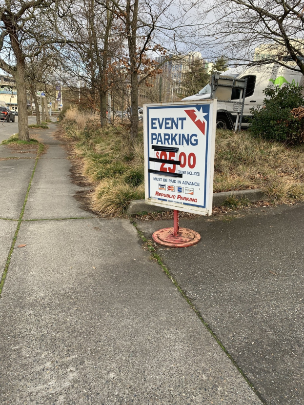 Schild am Straßenrand mit der Aufschrift "Event Parking" vor Gras, Pflanzen, Bäumen, Fahrzeugen, Gebäuden und einem klaren blauen Himmel.