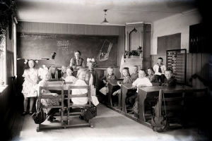 Schwarzes und weißes Foto einer Klasse mit Kindern auf Bänken sitzend und stehend, Taschen auf Bänken, eine Tafel, Topfpflanzen, ein Schrank, ein Fenster und Deckenleuchten.