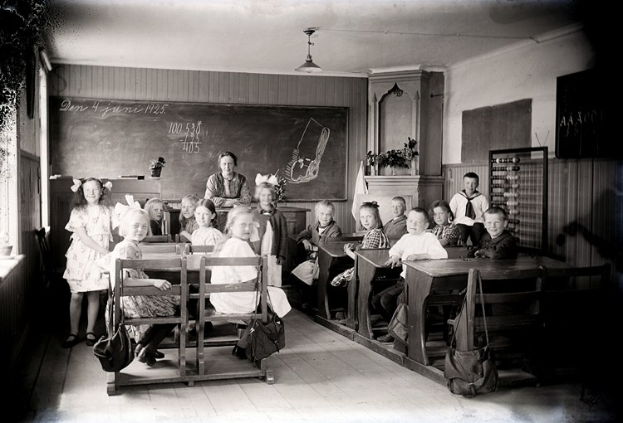 Schwarzes und weißes Foto einer Klasse mit Kindern auf Bänken sitzend und stehend, Taschen auf Bänken, eine Tafel, Topfpflanzen, ein Schrank, ein Fenster und Deckenleuchten.