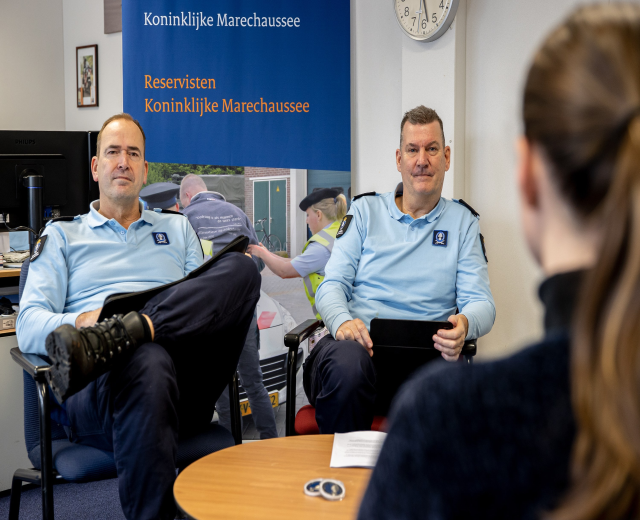 Zwei Polizeibeamte sitzen an einem Tisch mit Papieren und Gegenständen und sprechen mit einer Frau, im Hintergrund eine Wand mit einem Banner, einer Uhr und Foto Rahmen, sowie ein weiterer Tisch mit einem Monitor, Tastatur und Maus links.
