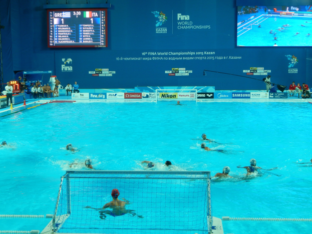 Wasserballspieler bei den 2016 FINA-Weltmeisterschaften in Kazan im Pool gegeneinander antretend, mit Zuschauern auf der linken Seite und einem Anzeigeboard mit Event-Branding im Hintergrund.
