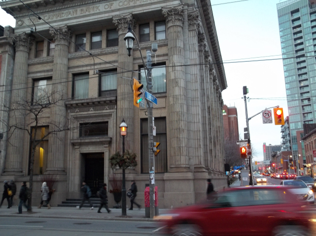 Eine belebte Stadtstraße mit Fahrzeugen, Fußgängern, Verkehrszeichen, Schildern, Bäumen und Gebäuden unter einem sichtbaren Himmel, mit einem roten Auto im Vordergrund und einem großen Gebäude.