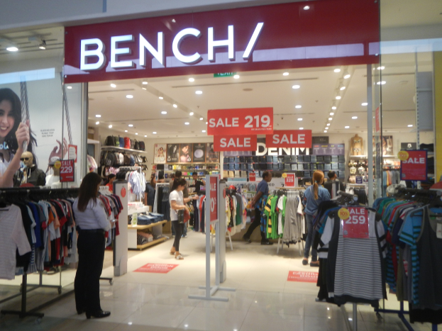 Ein Bekleidungsgeschäft namens "Bench" in einem Einkaufszentrum mit hängenden Kleidern, Informationsschildern, Deckenleuchten und einem Bildschirm auf der linken Seite.