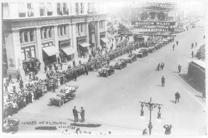Ein altes Schwarz-Weiß-Foto eines Umzugs auf einer Straße mit Menschen, Fahrzeugen, Laternenmästen, einem Schild, Gebäuden und einem bewölktem Himmel.