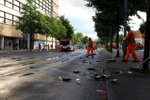 Eine Gruppe von Menschen in orangefarbenen Uniformen reinigt Straßenmüll von einer Straße, mit verstreuter Müll, Bäumen, Polen, Brettern, Fahrzeugen und bewölktem Himmel im Hintergrund.