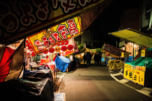 Ein belebter Nachtmarkt mit zahlreichen beleuchteten Ständen und Menschen, die eine von Gebäuden gesäumte Straße entlanggehen, mit einer Person im Vordergrund.