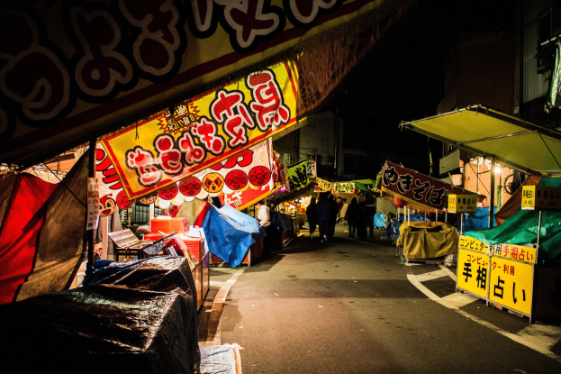Ein belebter Nachtmarkt mit zahlreichen beleuchteten Ständen und Menschen, die eine von Gebäuden gesäumte Straße entlanggehen, mit einer Person im Vordergrund.