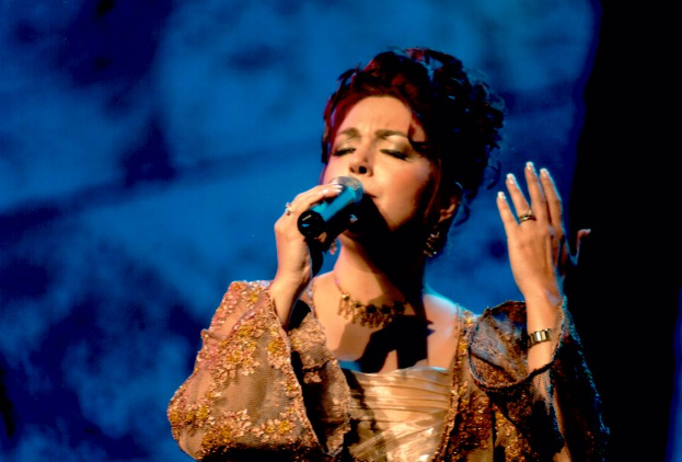 Eine Frau in einem goldenen Kleid singt in ein Mikrofon auf einer Bühne mit einem blauen und schwarzen Hintergrund und performt bei der Eurovision Song Contest.