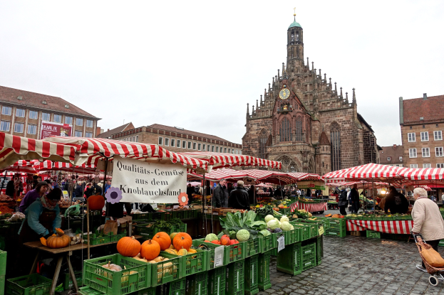 Ein belebter Markt in Nürnberg, Deutschland, mit Ständen, die verschiedene Früchte und Gemüse anbieten, Menschen, die einkaufen und umhergehen, und Zelten, die um den Bereich herum aufgebaut sind, mit Gebäuden und einem Uhrenturm im Hintergrund unter einem sichtbaren Himmel.