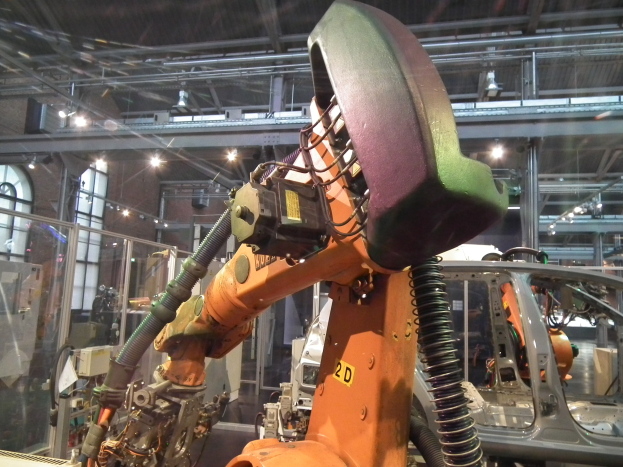 Roboterarm bei der Montage eines Autos in einer industriellen Fabrikumgebung mit Metallstangen, Beleuchtung und einer Glaswand auf der linken Seite.