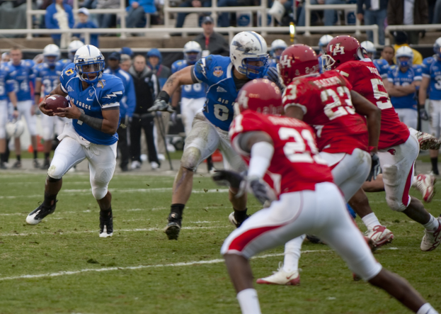 Ein Fußballspieler in Uniform rennt mit dem Ball, während er von Gegenspielern in Helmen und Sportbekleidung verfolgt wird, mit Zuschauern und Geländern im Hintergrund auf einem Rasenfeld.