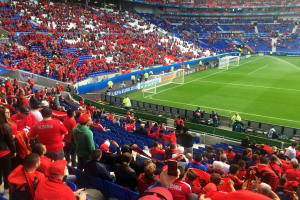Ein volles Stadion mit Zuschauern, einige sitzen, andere stehen, schauen ein Fussballspiel, mit Bannern, einem Tor und Deckenbeleuchtung.