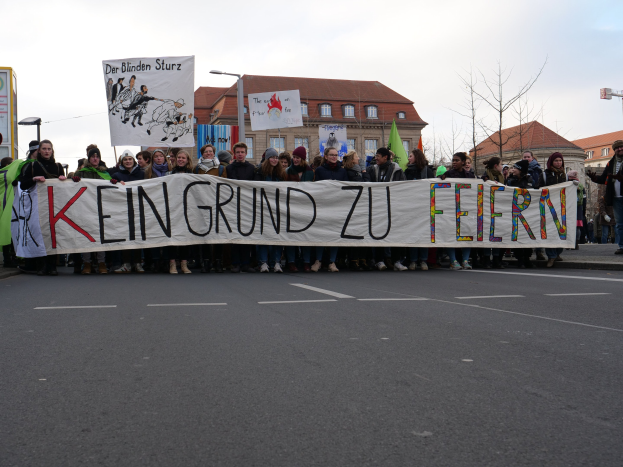 Demonstranten mit einem Banner mit der Aufschrift "Kein Grund zu Feiern" gegen deutsche Sparmaßnahmen, mit Gebäuden, Bäumen und einem klaren Himmel im Hintergrund.