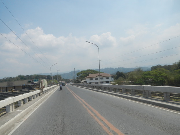 Ein Motorradfahrer fährt auf einer von Leitplanken gesäumten Autobahn, flankiert von Laternen, Gebäuden mit Fenstern und Bäumen, mit Hügeln und einem bewölkten Himmel im Hintergrund.