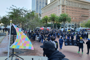 Eine große Gruppe von Menschen steht vor einer Menge, die Fahnen und Banner schwingt, mit einem Redner, Bäumen, Zelten, Laternenmasten und Gebäuden im Hintergrund, wahrscheinlich beim San Francisco LGBT Pride Parade.