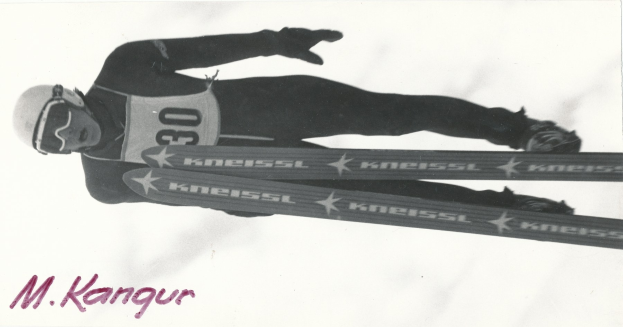 Schwarzes und weißes Foto eines Skifahrers in der Luft, der einen Helm, eine Brille, Handschuhe und Schuhe trägt und Skiplatten hält, mit Text unten.