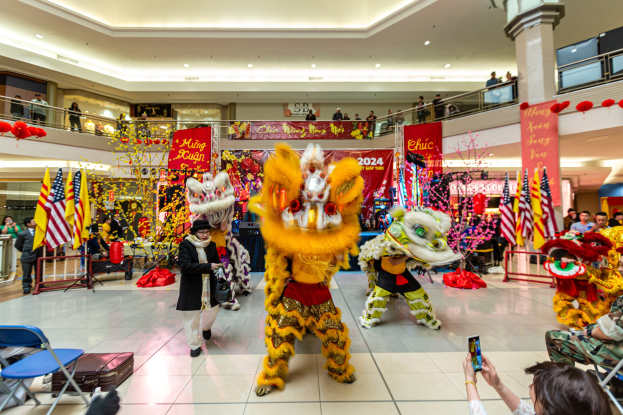 Eine lebendige Feier des chinesischen Neujahrs in einem Einkaufszentrum mit tanzenden Menschen in Kostümen, anderen, die auf Stühlen sitzen und Handys verwenden, umgeben von Fahnen, Bannern, Dekorationen, Geländern, Säulen und Deckenlampen, mit einem Wasserzeichen unten.