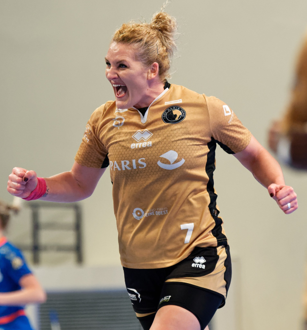 Eine Frau in brauner Bluse und schwarzen Shorts rennt auf einem Futsalplatz, feiernd mit triumphaler Miene, während zwei andere Frauen im Hintergrund zu sehen sind.