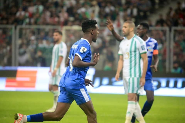 Ein Fussballspieler in einem blauen und weissen Trikot feiert mit erhobenen Armen, umgeben von einer Gruppe in weissen und grünen Trikots, mit einem unscharfen Hintergrund, der ein weisses und blaues Board und ein Geländer zeigt.