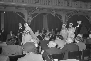 Ein Schwarz-Weiß-Foto einer Hochzeitszeremonie in einem großen Raum mit vielen Gästen, die sitzen und stehen, einige halten Blumensträuße, und Tische mit Tassen und anderen Gegenständen, mit Vorhängen und Deckenlampen im Hintergrund.