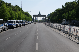 Eine Reihe von Polizeiwagen auf einer Straße vor dem Brandenburger Tor in Berlin, Deutschland, mit Menschen auf Fahrrädern und in der Nähe Stehenden, Absperrungen, Bäumen und einem Bogen mit Statuen im Hintergrund.