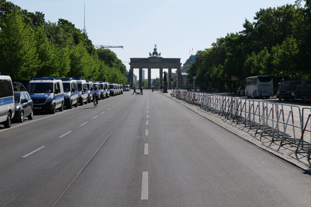 Eine Reihe von Polizeiwagen auf einer Straße vor dem Brandenburger Tor in Berlin, Deutschland, mit Menschen auf Fahrrädern und in der Nähe Stehenden, Absperrungen, Bäumen und einem Bogen mit Statuen im Hintergrund.