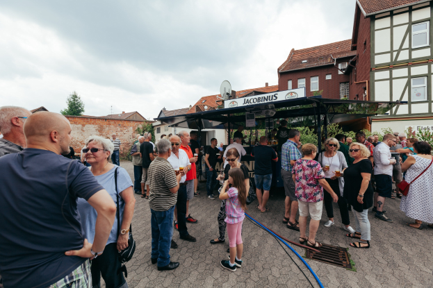 Gruppe von Menschen auf einem Outdoor-Bierfest vor einem Gebäude mit Fenstern, umgeben von Bäumen unter einem bewölkten Himmel, mit einer Hütte mit einem Namensschild im Hintergrund.