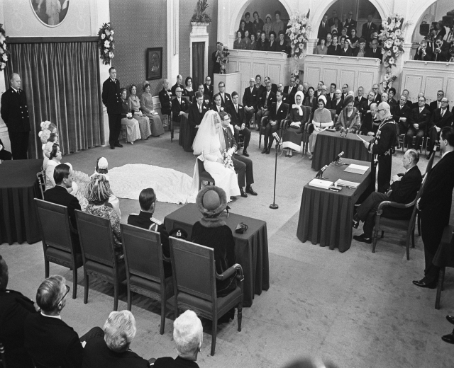 Schwarzes und weißes Foto einer Hochzeitszeremonie in einem großen Raum mit sitzenden und stehenden Gästen, Tischen mit Mikrofonen und Papieren, einer Wand mit Rahmenfotos, Blumenbouquets, einem Vorhang und einem Podium mit Mikrofonständer.