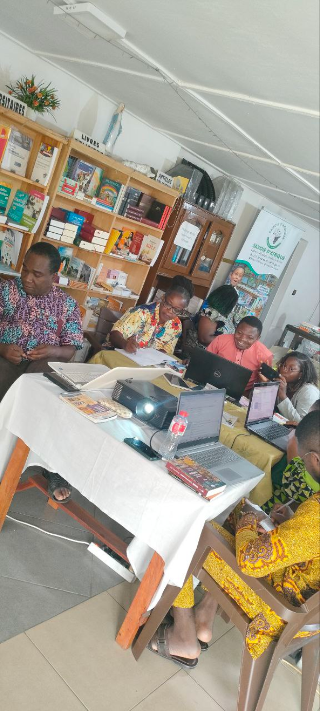 Eine Gruppe von Menschen sitzt um einen Tisch mit Laptops, Büchern, einem Projektor und anderen Gegenständen, mit Bücherregalen und einer Blumenvase im Hintergrund und einem Banner, das eine Workshop-Veranstaltung zum Thema Bildung in Ghana ankündigt.
