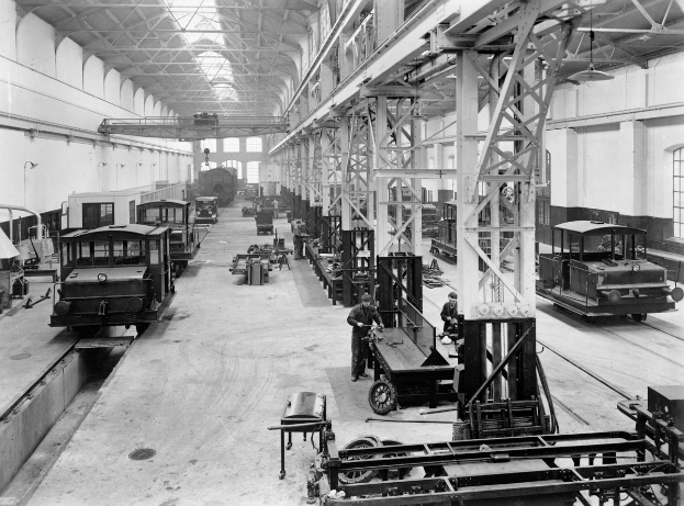 Schwarzes und weißes Foto einer Fabrikhalle mit mehreren Bahnen unter Reparatur, Arbeitern, die sich darum kümmern, Metallpfosten, die die Struktur stützen, Deckenlampen und Fenstern an den Wänden.