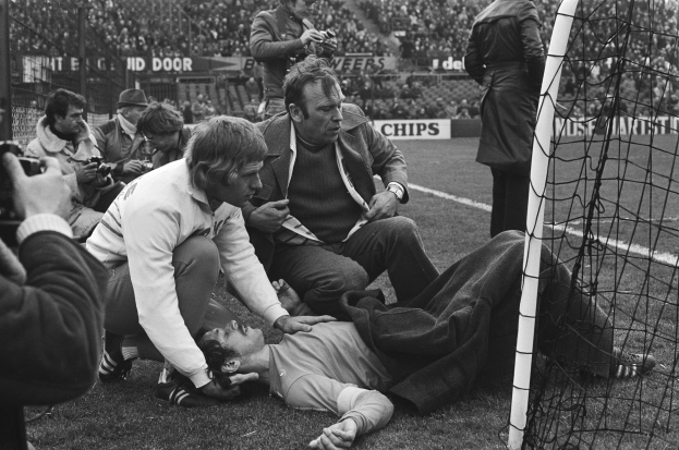 Ein Mann liegt in der Nähe eines Fußballtors auf dem Boden, während eine Gruppe von Menschen auf dem Rasen um ihn herum sitzt und steht, mit Zuschauern auf den Tribünen und Informationsschildern im Hintergrund.