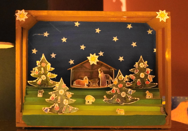 Eine Krippe des Heiligen Abends in einer Holzkiste auf einem Tisch, mit einer Lampe links und einer Wand im Hintergrund, geschmückt mit Weihnachtsbäumen, Tieren und einem Haus.