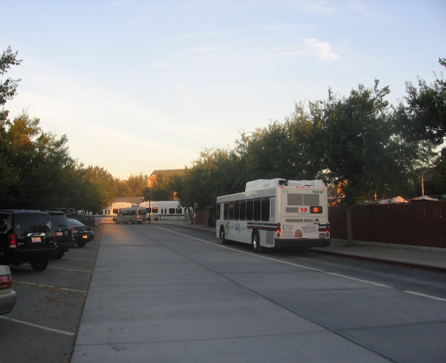 Ein Shuttle-Bus fährt auf einer Straße mit parkenden Autos, flankiert von Bäumen und Gebäuden unter einem klaren blauen Himmel.