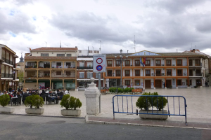 Ein belebter Stadtplatz mit Menschen auf Stühlen sitzend und stehend, umgeben von Topfpflanzen, Metallabsperrungen, Straßenlaternen mit Flaggen, einem Schild, Gebäuden und einem bewölkten Himmel.