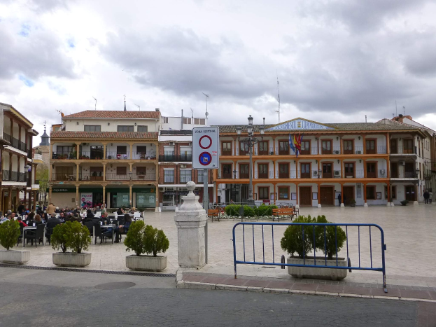 Ein belebter Stadtplatz mit Menschen auf Stühlen sitzend und stehend, umgeben von Topfpflanzen, Metallabsperrungen, Straßenlaternen mit Flaggen, einem Schild, Gebäuden und einem bewölkten Himmel.