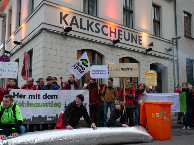 Eine Gruppe von Menschen mit Protestschildern und Transparenten vor einem Gebäude in Deutschland, mit zwei Personen im Vordergrund und einem Müllcontainer rechts.