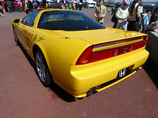 Ein gelber Sportwagen vor einer Menge, einige tragen Helme, mit einer Wand rechts und Zelten, Bäumen, Gebäuden und einem klaren blauen Himmel im Hintergrund.