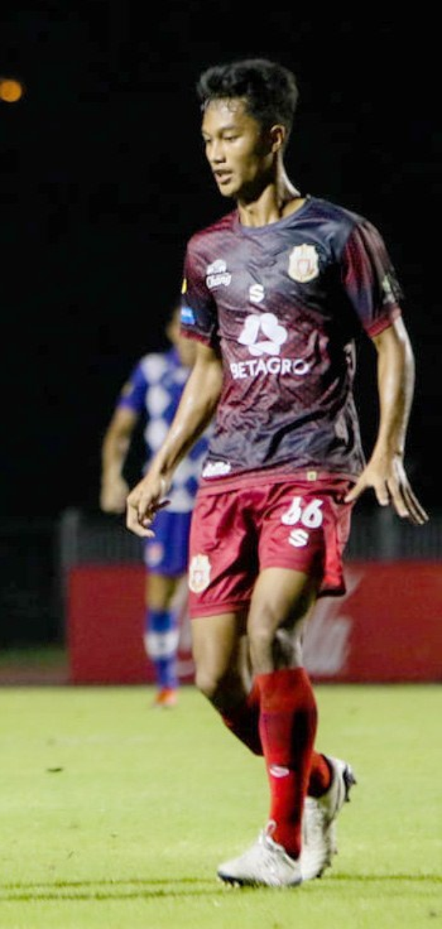 Ein Mann in einem rotbraunen Hemd und roten Shorts spielt Fußball auf einem Feld, mit einer anderen Person im Hintergrund und einer Tafel mit Text.