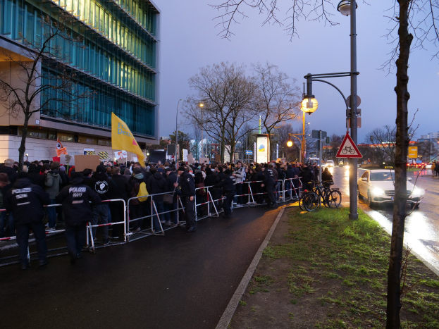 Eine große Gruppe von Menschen mit Schildern steht vor einem Gebäude mit Barrikaden, Fahrrädern, Laternenmasten, Schildern, Bäumen und Gras unter einem sichtbaren Himmel, was auf eine Protestaktion in Berlin hinweist.